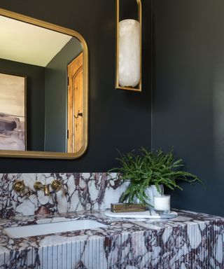 powder room with dark olive green walls, marble countertops and brass mirror and wall sconce