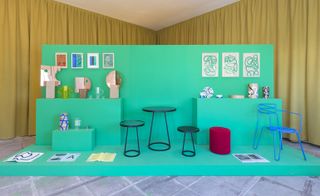 Interior set displayed against a green background, brass mirrors and wood vases etched with abstract drawings, thrfee little black stools, blue wire chair, mustard yeloow curtain backdrop, grey checked floor