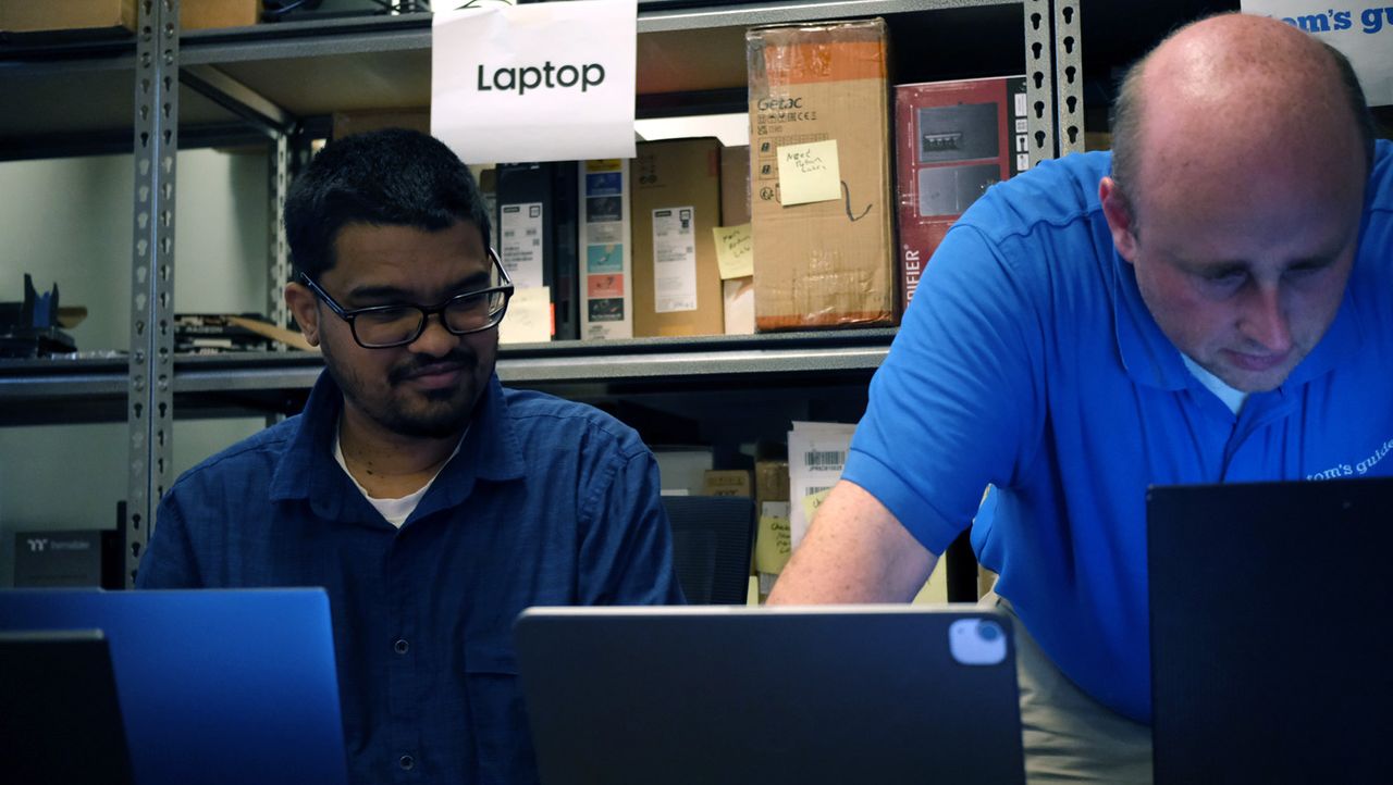 One person leans over a desk, another sits beside, while they both look at a desk with several laptops on top.