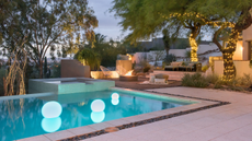 A luxury garden with a pool with floating orb lights, trees wrapped in fairy lights, and a firepit in the corner