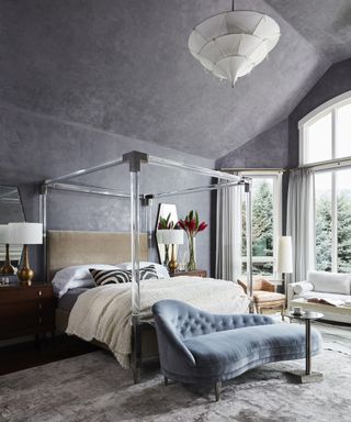 A bedroom with grey-blue textured plaster on the walls and ceiling, mirrored four poster bed frame, blue chaise at foot of the bed, and a seating area with white sofa in the window