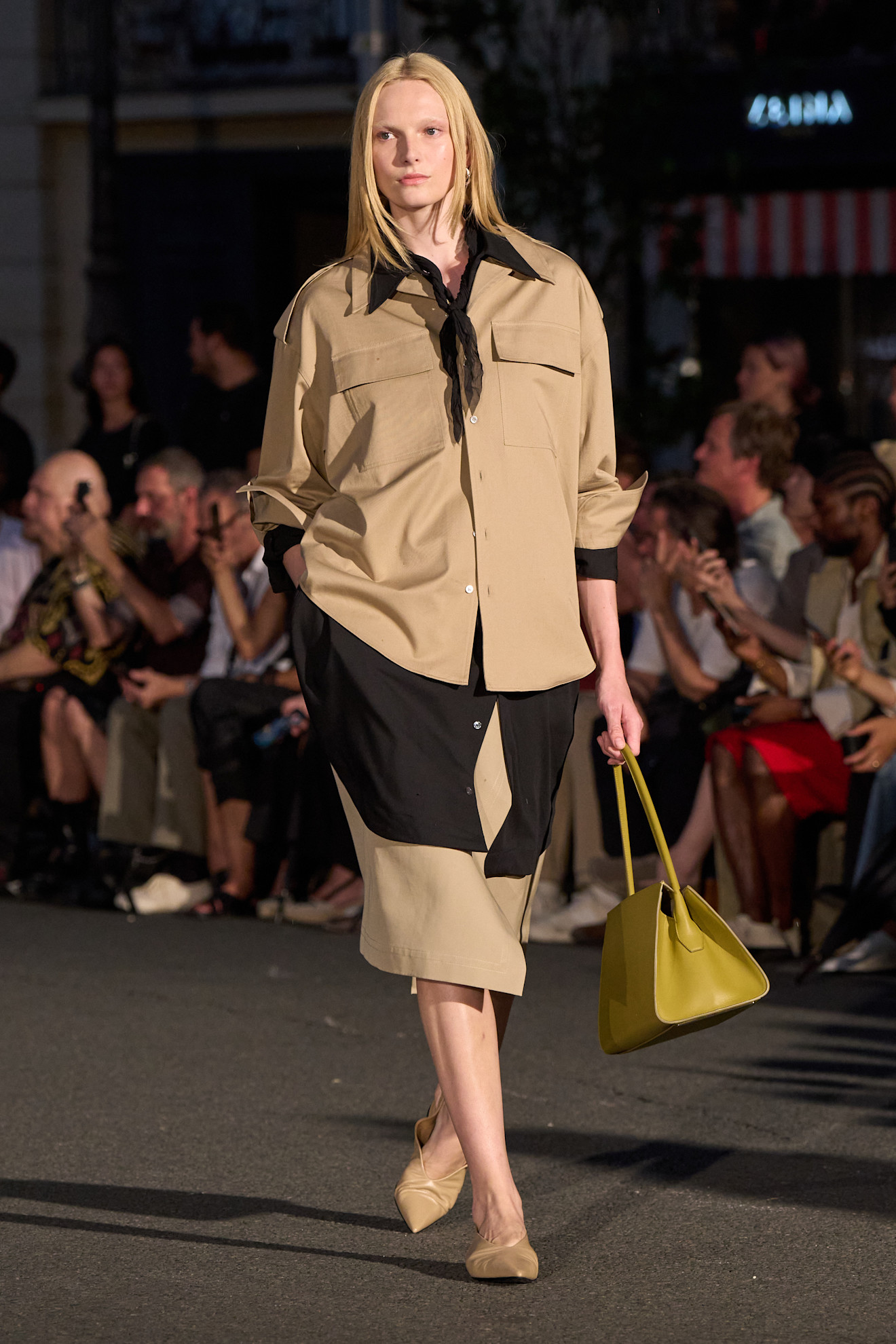 Layering staples for spring 2026 are shown in a photo from Ami's runway show, with a model walking while wearing an oversized tan shirt layered ontop of a black button-down shirt styled with a tan skirt, tan pumps, and a yellow handbag