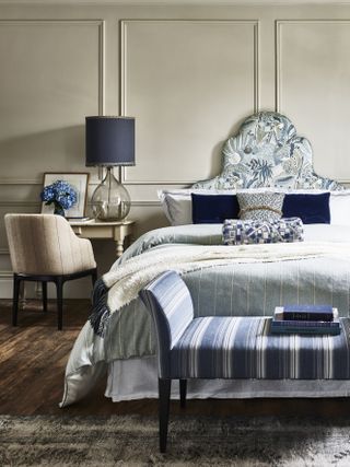blue and white bedroom with taupe walls, blue and white bedding, footstool, wooden floors, blue lamp, armchair, side table