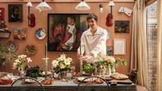 Man in a white shirt places a green candle on a decorated table surrounded by flowers, art-covered peach walls, and a cat watching.