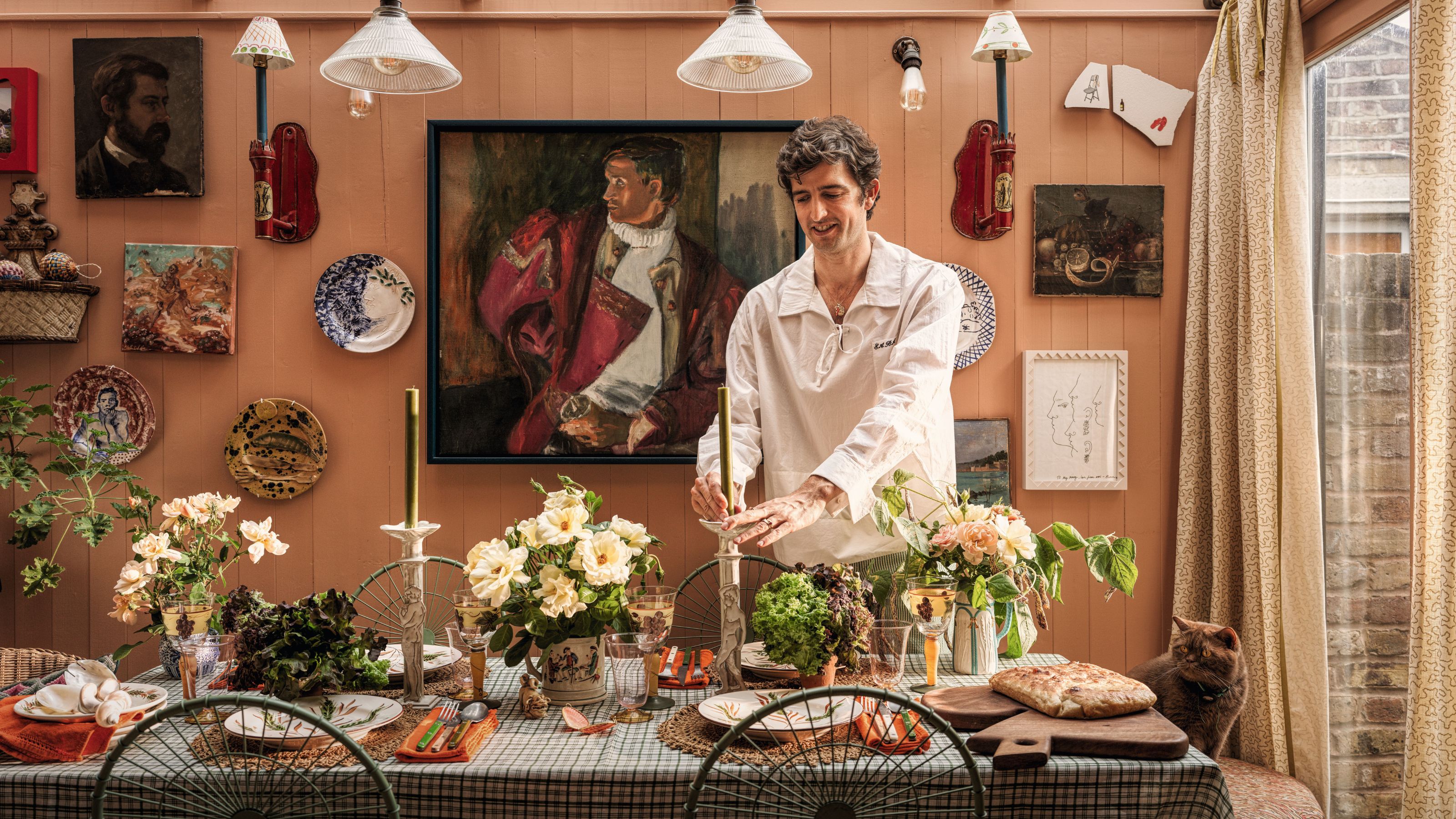 Man in a white shirt places a green candle on a decorated table surrounded by flowers, art-covered peach walls, and a cat watching.