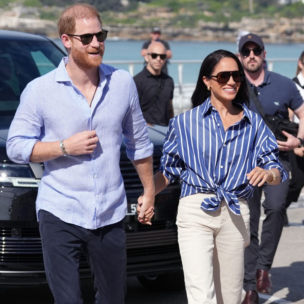 Meghan Markle holds hands with Prince Harry wearing white cropped jeans with white trainers and a blue striped shirt.