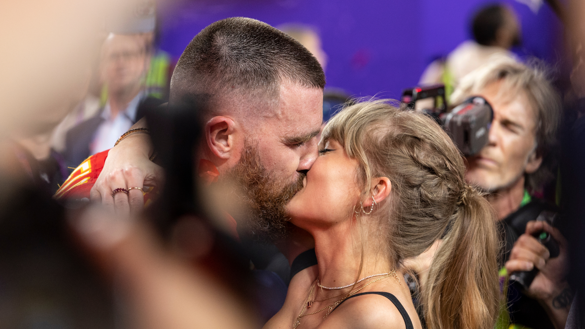 Taylor Swift has a ponytail and red lipstick and kisses NFL football player Travis Kelce as cameras film them