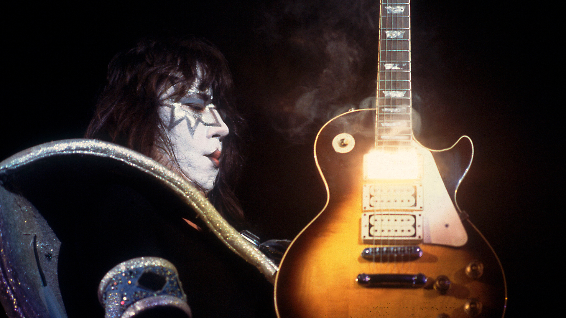 A man wearing black and white make up looks at a glossy brown electric guitar