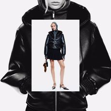 The model wears a black faux leather hooded jacket with a zipper, gray quarter zip inside the hoodie, and a pleated mini skirt with studded pointed-toe heels