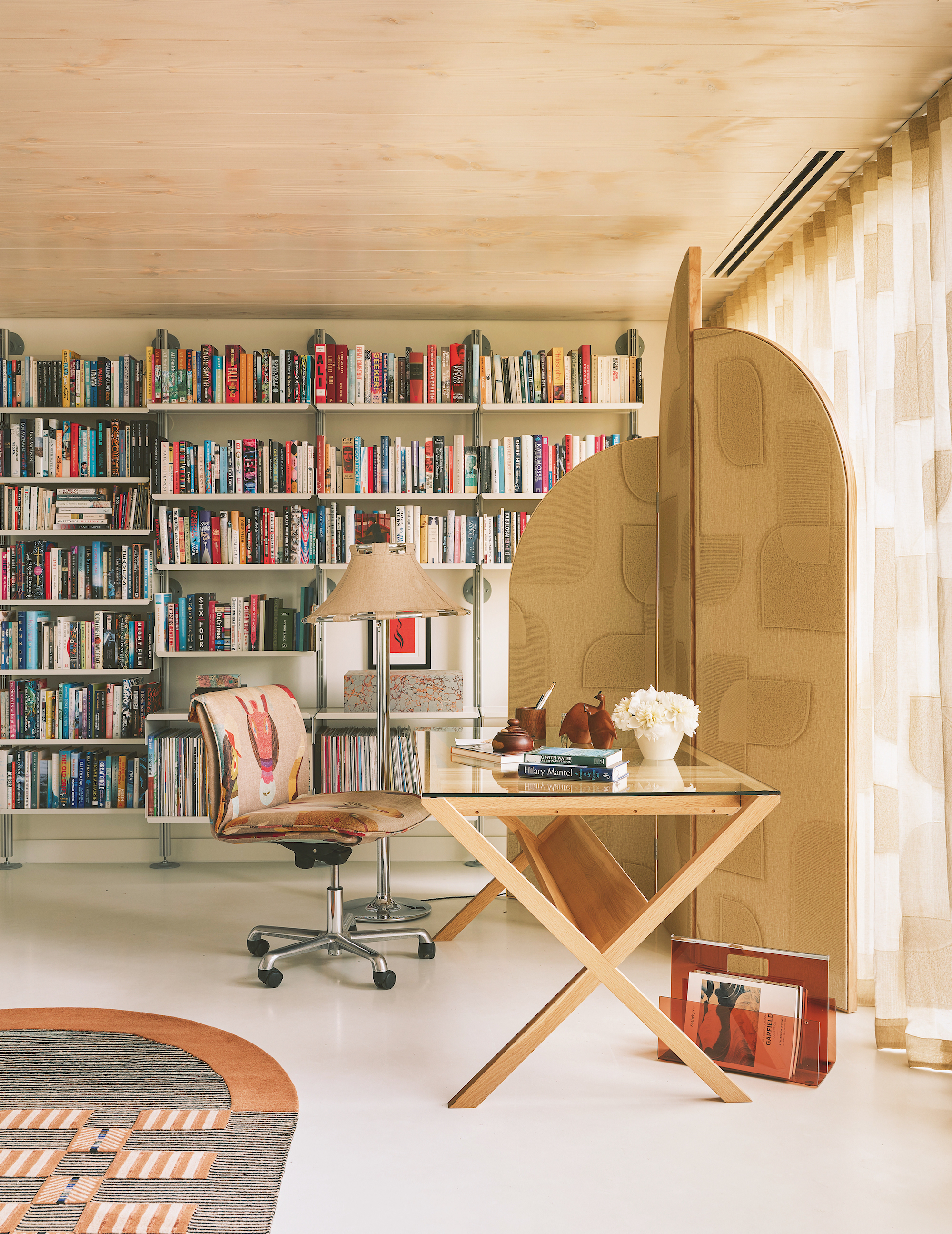 A modern study with a glass desk facing with window, with a matching patterned partition and curtains. There is a floor to ceiling book shelf on the far wall and a rug in the left hand corner.