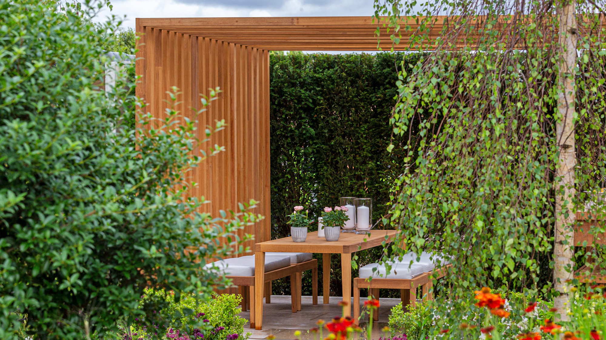Modern slatted pergola design with dining area and garden furniture, a tree, shrubs and flowers