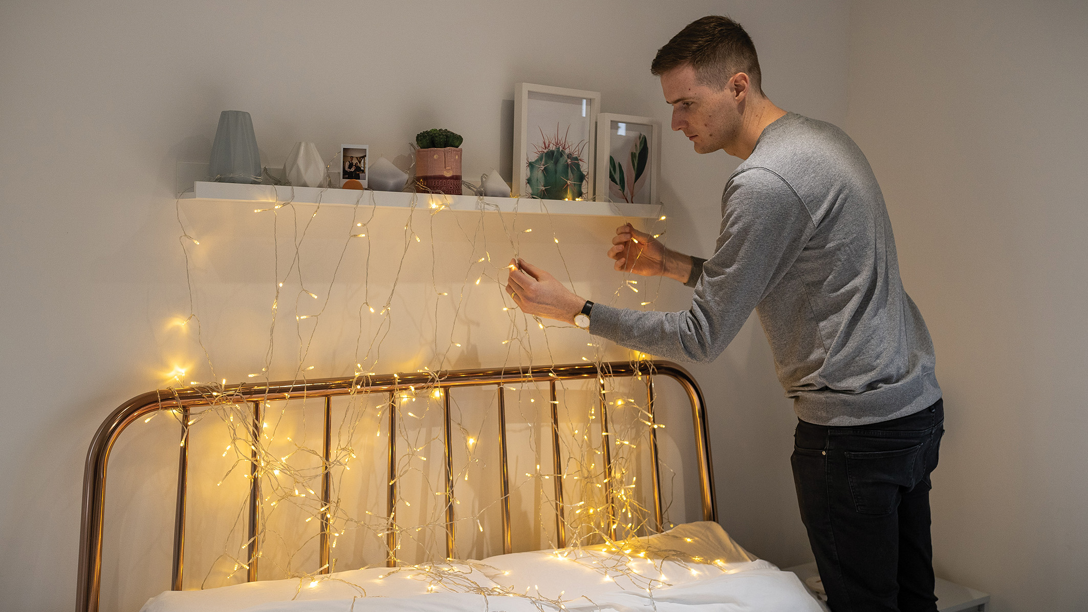 A person decorates a bedroom with warm fairy lights above a metal bed frame. The room has a cozy, modern feel with plants and framed art on a shelf