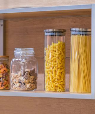 A wooden shelf with two mason jars and two cylinder storage containers filled with yellow pasta, biscuits, and colorful dried fruit