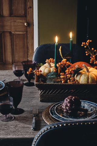 Halloween table decor with basket full of gourds, berries and green candles, blue patterned plates with rattan placemats, berry coloured glassware