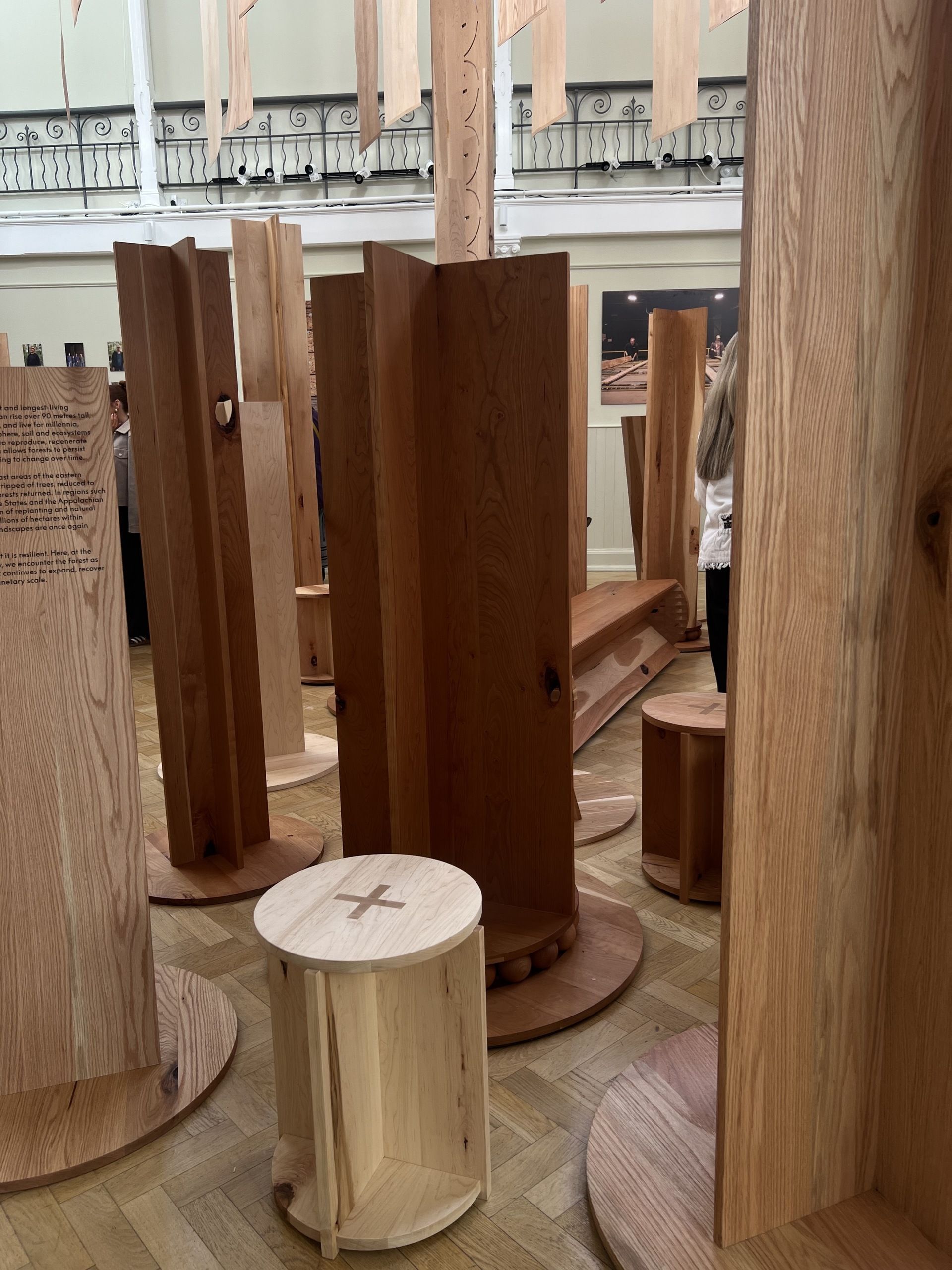 Image of a gallery space with wooden forms arranged throughout the space. There are also wooden stools mixed in.