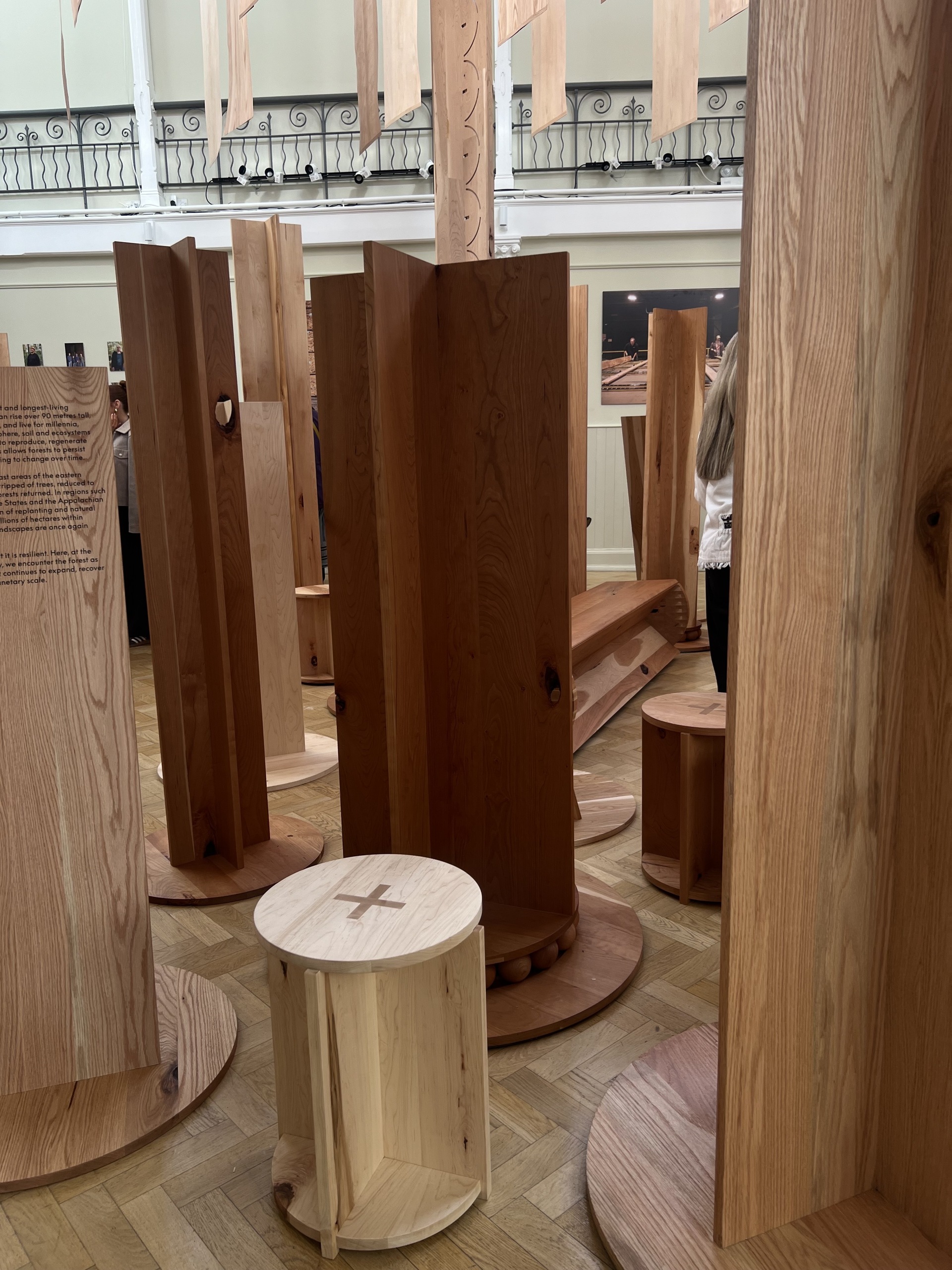 Image of a gallery space with wooden forms arranged throughout the space. There are also wooden stools mixed in.