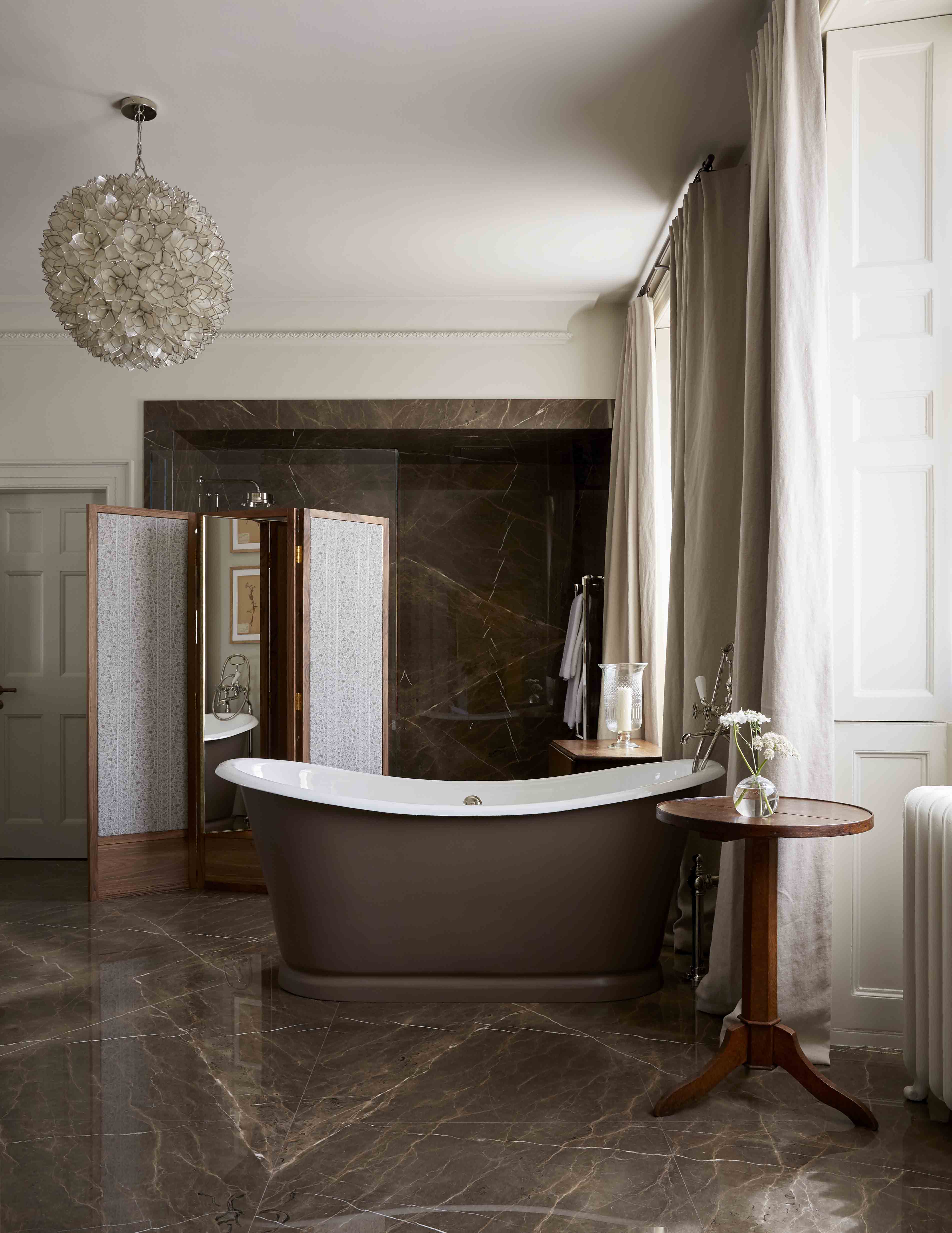A transitional style hotel bedroom features a sleek, large bathtub, brown marble floors, wood and mirror room dividers, and plush curtains.