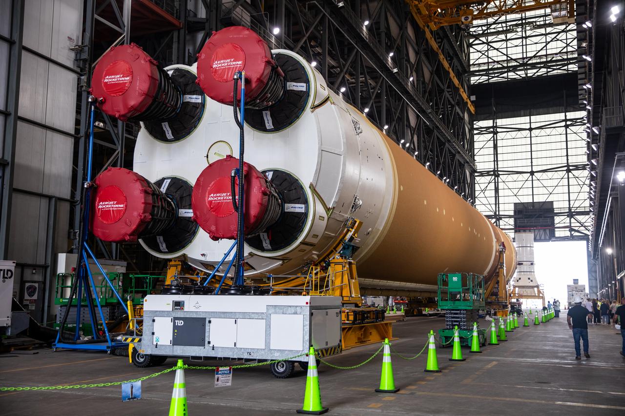 a rocket piece on its side with engines facing the camera. the engines are covered in protective plastic. the rocket is inside a large warehouse and surrounded by traffic cones