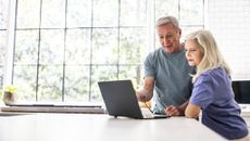 A senior couple standing in their kitchen, reviewing finances together on a laptop.