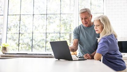 A senior couple standing in their kitchen, reviewing finances together on a laptop.