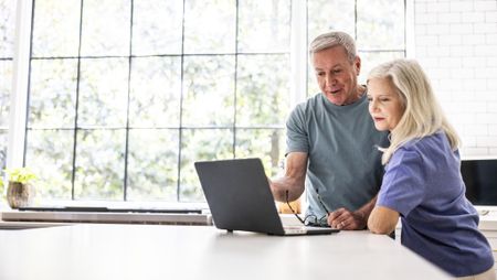 A senior couple standing in their kitchen, reviewing finances together on a laptop.
