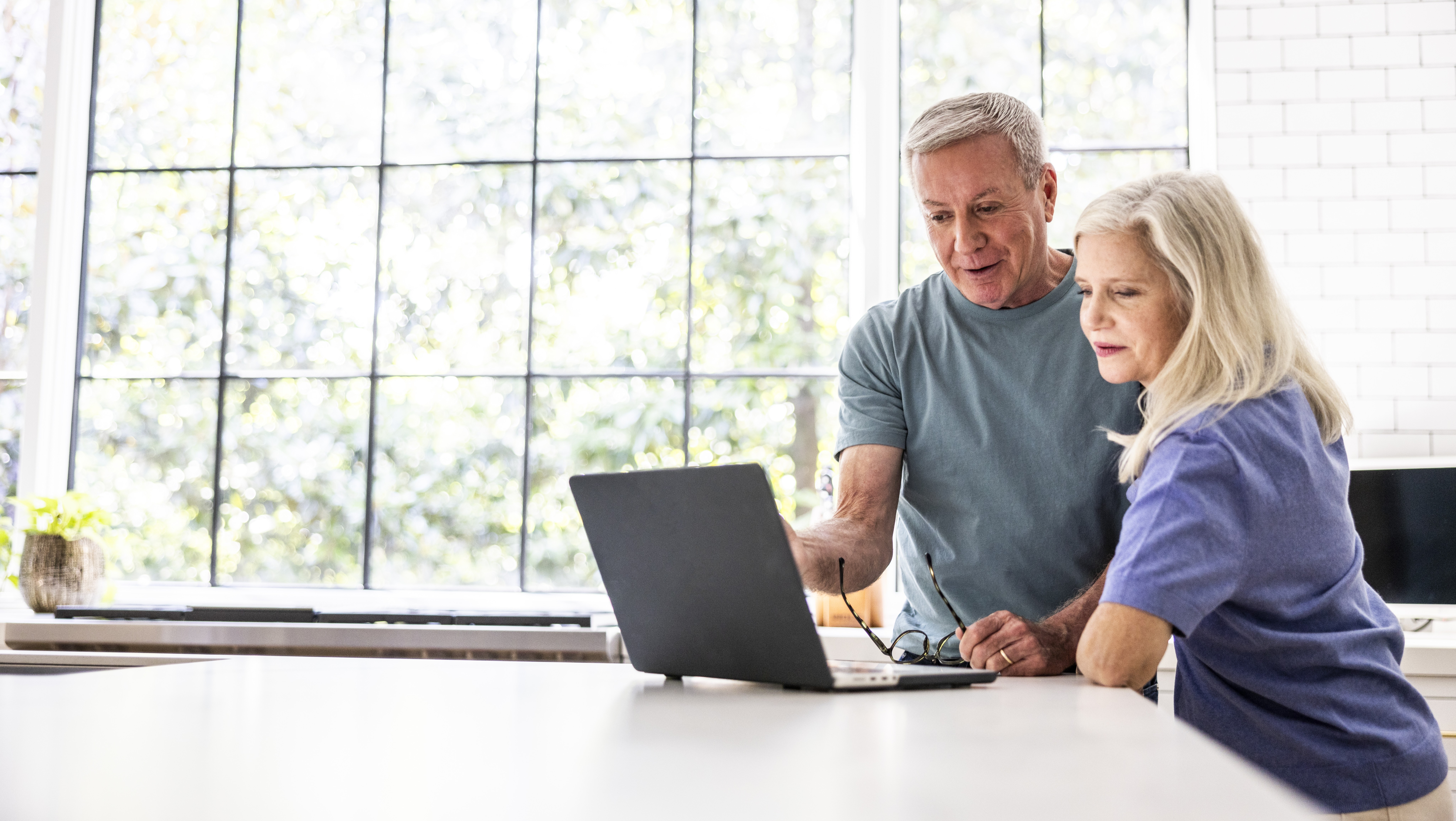 A senior couple standing in their kitchen, reviewing finances together on a laptop.
