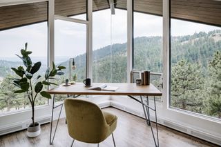 Wooden desk with yellow accent chair overlooking forest view out of picture windows