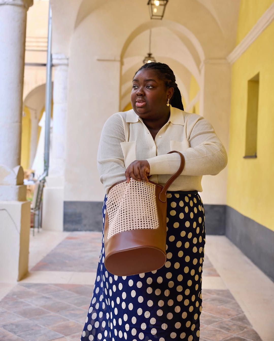 @abimarvel wears a blue-and-white polka dot skirt with a cream cardigan and raffia bucket bag