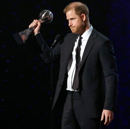 Prince Harry holding up the Pat Tillman Award for Service in a black suit and tie