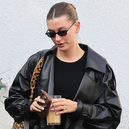 Hailey Bieber leaves pilates wearing a leather trench coat with a pink manicure and an iced coffee