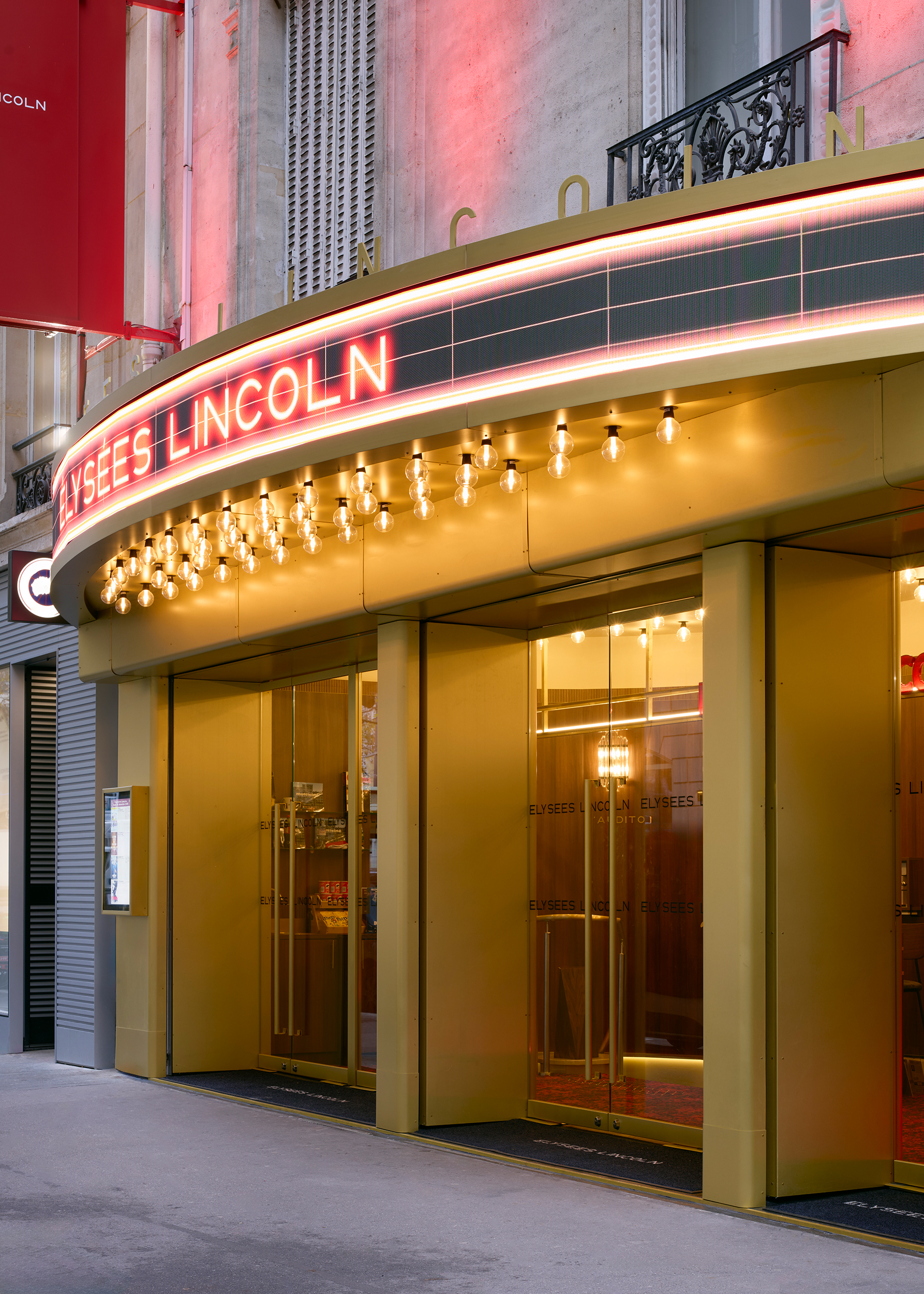 A series of interior shots depicting the retro-futuristic decor of a cinema captures velvet-clad rooms with a palette of reds, pinks, and pale dark greens, theatrical doorways, either arched or draped in curtains, ceiling porthole windows, and a vintage, golden entrance with red neon lights.