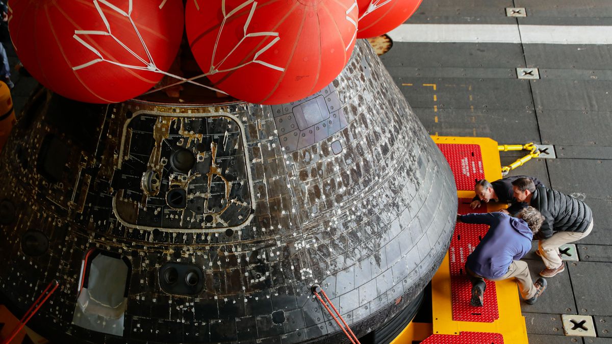 three technicians working alongside the orion spacecraft on a ship. the spacecraft has three inflatable devices on top