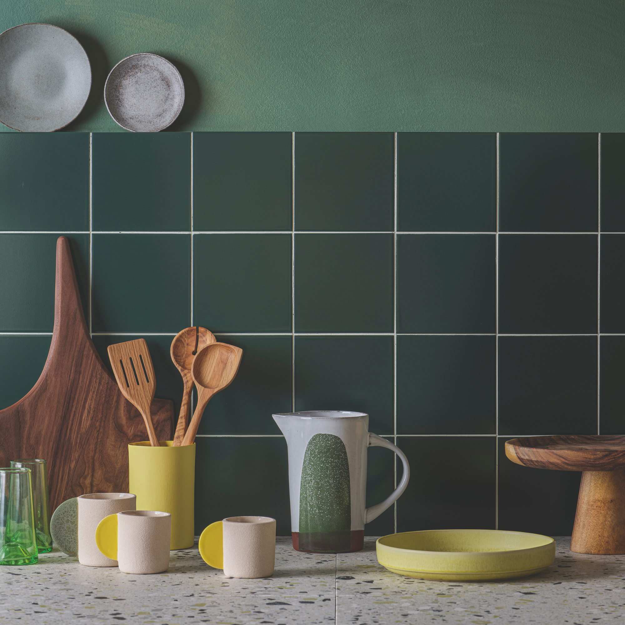 A blue and green kettle on a grey worksurface, There are three pink and yellow mugs next it, a yellow bowl and green tiling on the wall.