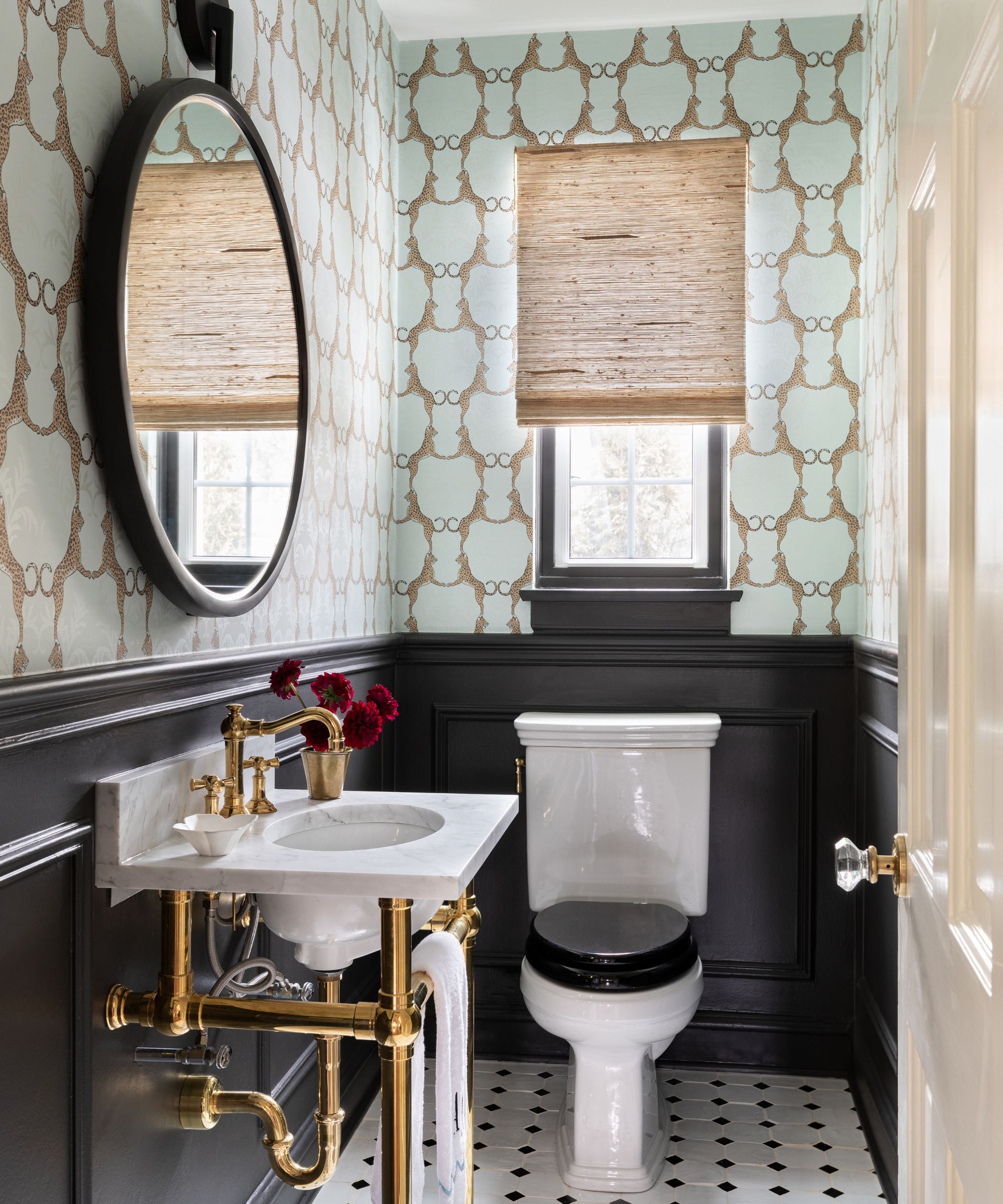 A traditional bathroom with black lower walls, aqua blue wallpaper on the upper walls, a marble sink with brass fixtures, and a round mirror above it.