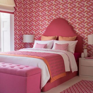 Vibrant pink bedroom, modern geometric wallpaper, large pink double bed, padded chest storage, pink and white bedlinen, bedside table. Pub Orig