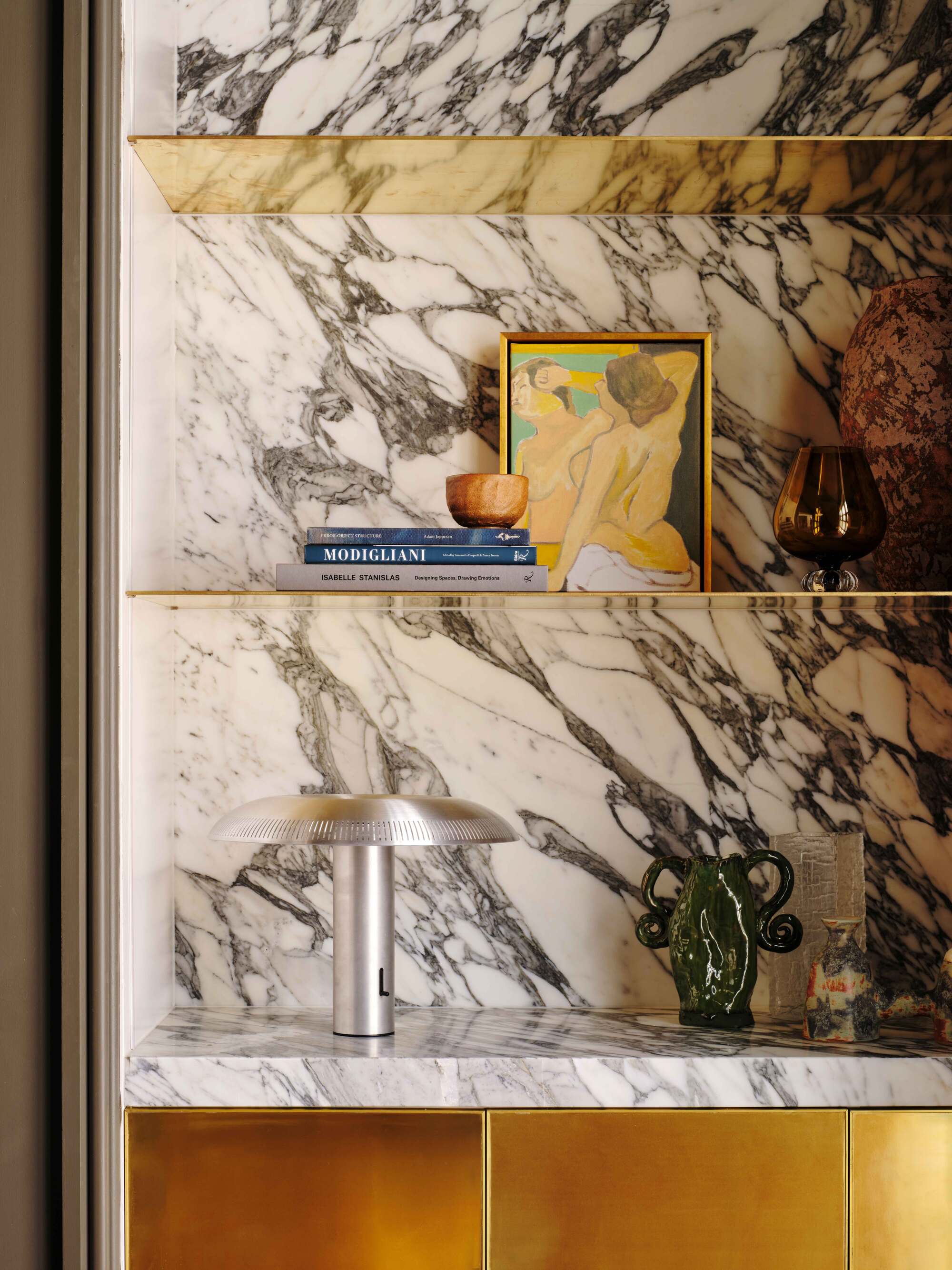 close up of marble bookcase with gold shelves styled with artwork, stack of books, silver lamp, and small ceramics