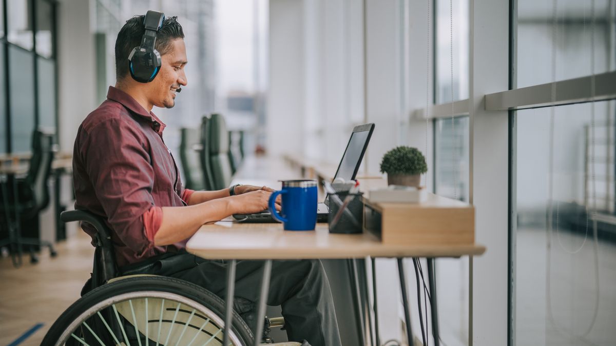 A South Asian disabled tech worker in a wheelchair, working on a laptop with a coffee on the desk to his right. Decorative: The worker is shot in medium-wide, with the top of his wheelchair visible and a large, modern office space clearly visible around and behind him.