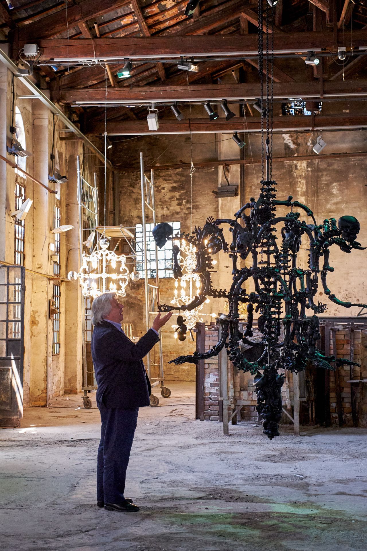 A middle-aged man wearing a dark suit stands in a glass workshop next to a monumental Murano glass chandelier shaped after human skulls and spines. Behind him, a warmly lit chandelier irradiates the scene.