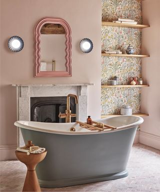 pale pink bathroom with fire place, roll top bath with gray exterior, floor tiles, modern wood stool, wallpaper in alcove with wood shelves, pink mirror, fluted wall lights