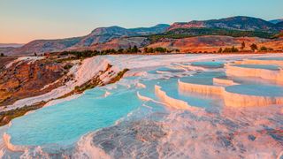 View of the Pamukkale travertines in orange-pink light. We see rocky mountains in the background and pools of spring water in the foreground.