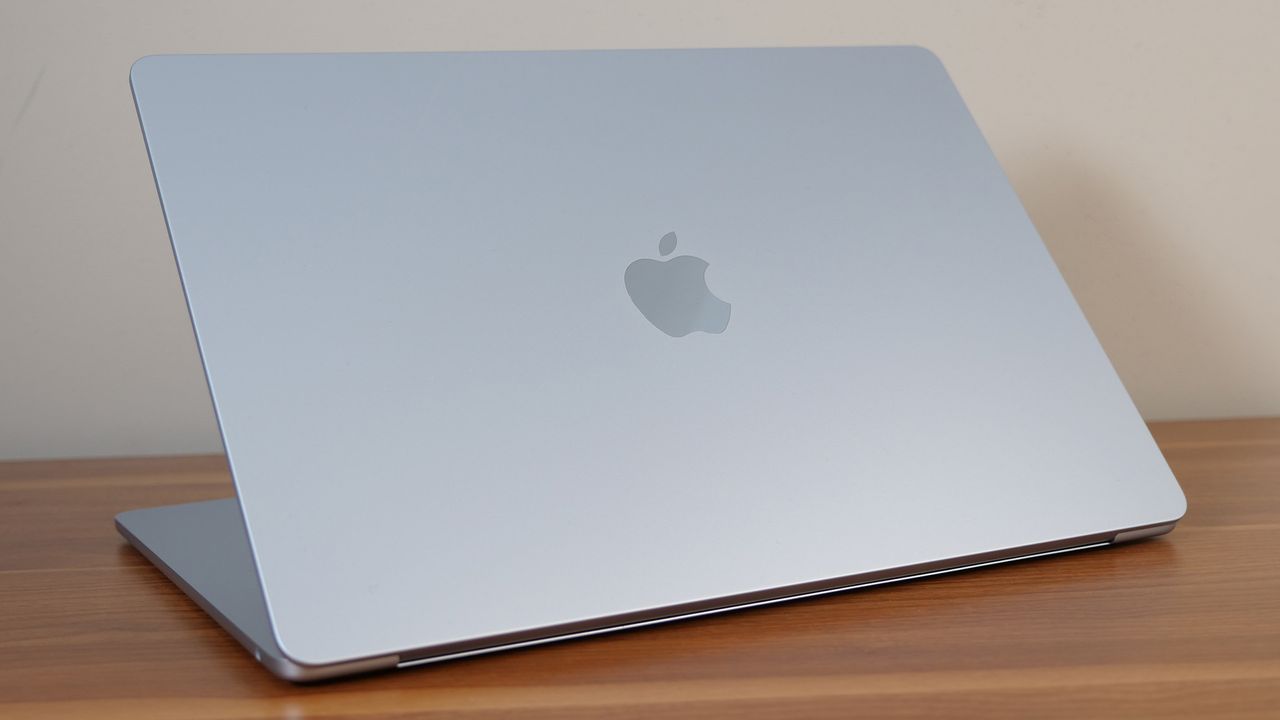 A silver laptop sitting on a wooden table at a slight angle with its lid half-open. A shinny, Apple logo is centered on the lid's surface.