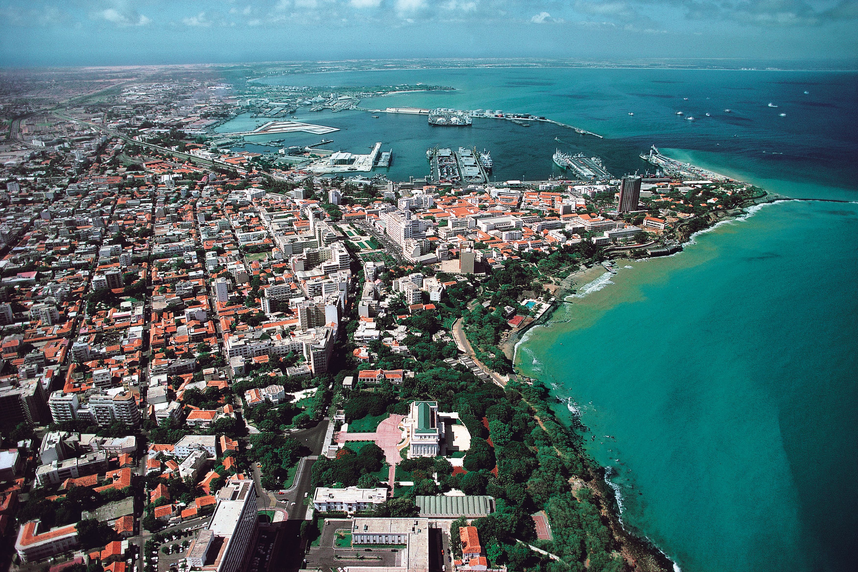 View of a seaside city from above, with lots of green areas and just as many roofs overlooking the coast.