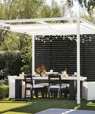 A green backyard area with a white pergola with black metal louvered panels, a dining table with glasses and flowers on it, and four black and white cushioned chairs, all on a grassy lawn