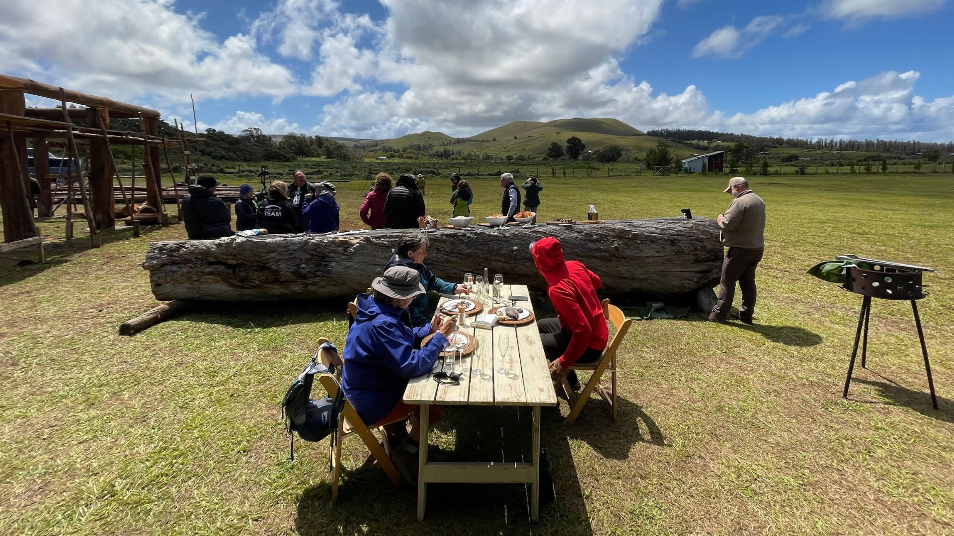 people sit around a long wooden table, while some stand in the background. the sky is blue and partly cloudy.