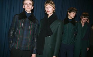 Four male models wearing looks from Paul Smith's collection. One model is wearing a blue high neck jumper, dark green, blue, black and red plaid style jacket with black short fur collar and black trousers. Next to him is a model wearing a dark green coat and black scarf. The third model is wearing a dark blue top, dark blue trousers and dark green jacket with black short fur collar. And the fourth model is wearing a green shirt and dark green and blue coat