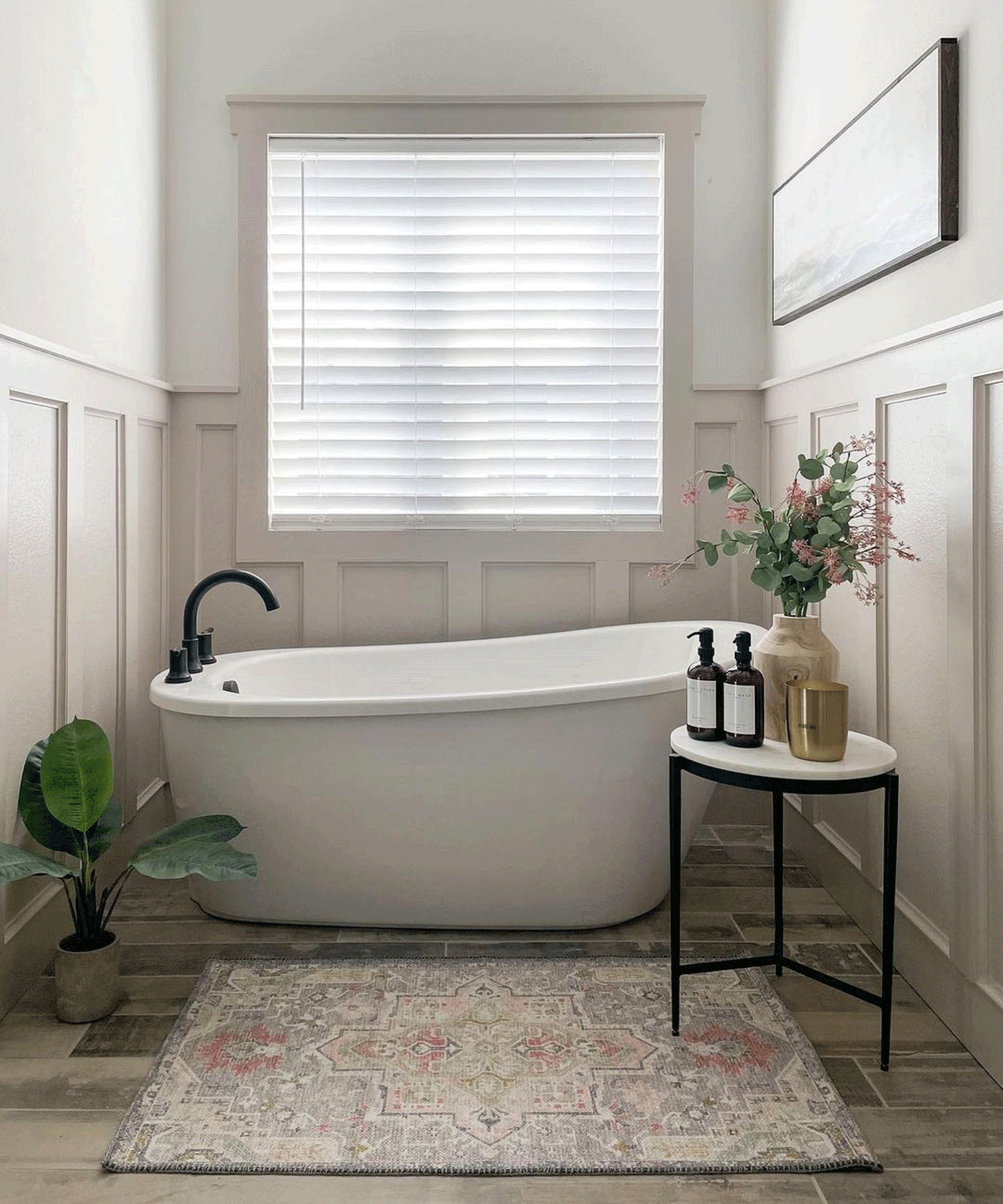 A small bathroom with bathtub, including blind window treatment, wall paneling (painted in a mid grey paint), side table, exotic rug and indoor houseplant