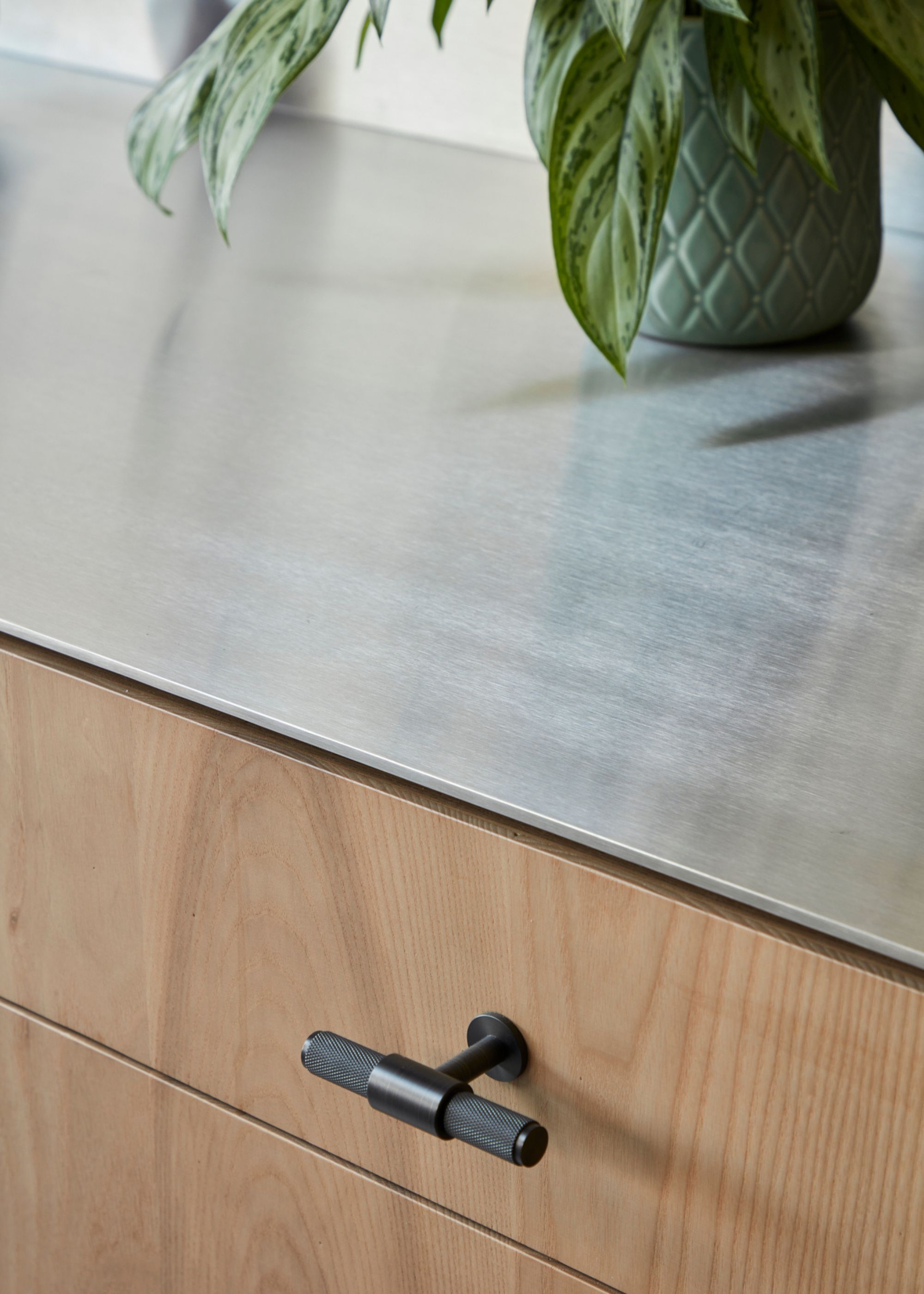 A close up shot of a stainless steel worktop in a kitchen with a view of one of the drawers with a handle. On top of the countertop is a cut off view of a plant in a pot