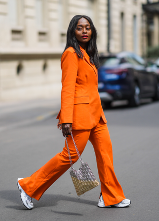 Carrole Sagba @linaose wears gold earrings, an orange belted Blazer jacket and matching suit flared Warehouse pants, white New balance sneakers, a shiny silver nailed Paco Rabanne handbag, on May 02, 2021 in Paris, France