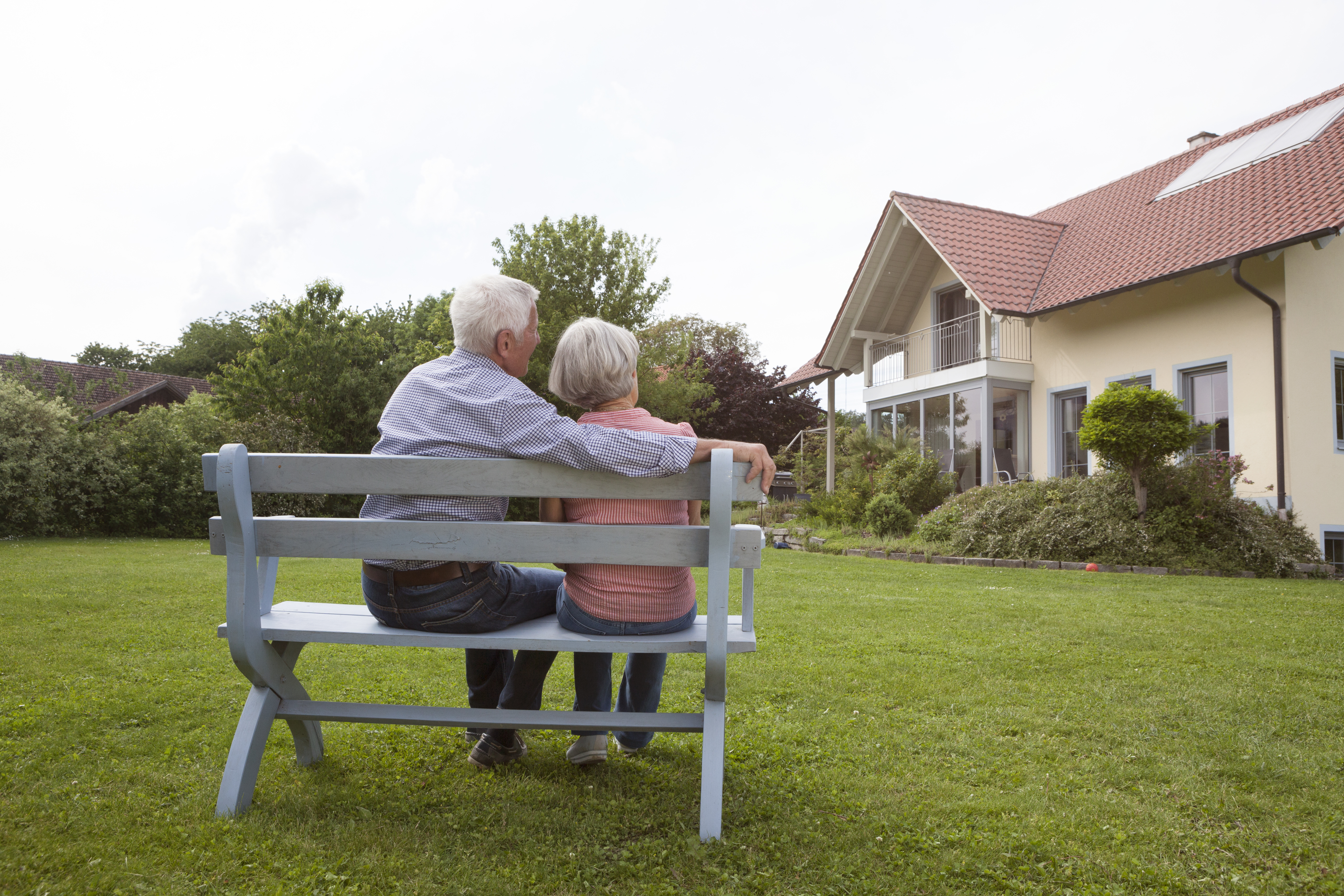 Pensioner couple sit on bench in the garden as they look at large property.
