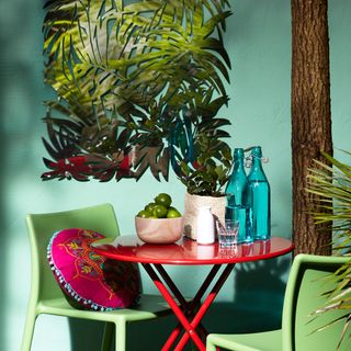 orange metal table and green chairs against a teal painted exterior wall and palm wall art
